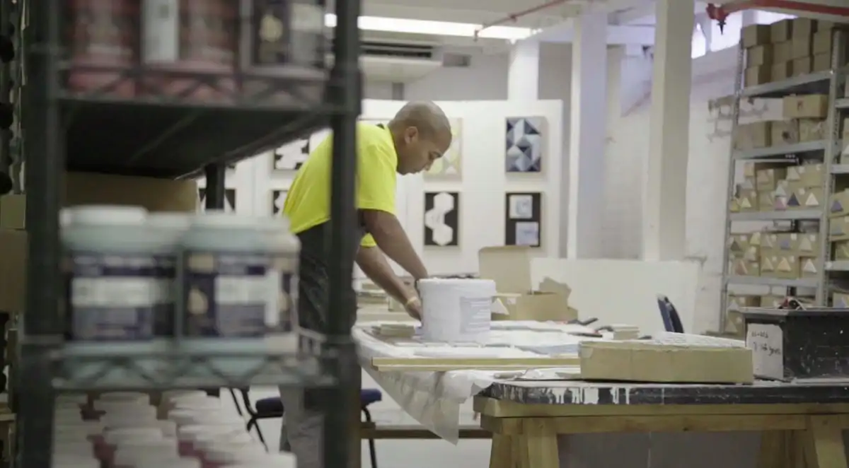 A person working on an art project in a studio, surrounded by shelves of supplies and artwork on the walls.
