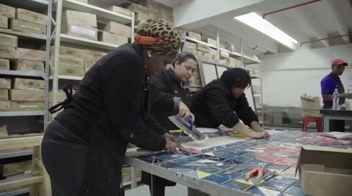 People working on colorful tile designs in a workshop, surrounded by shelves of boxes.