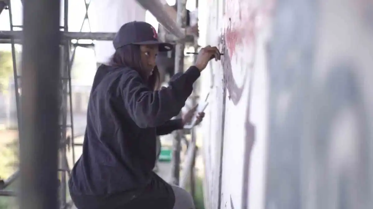 Person in a black jacket and cap spray-painting graffiti on a wall while standing on scaffolding.