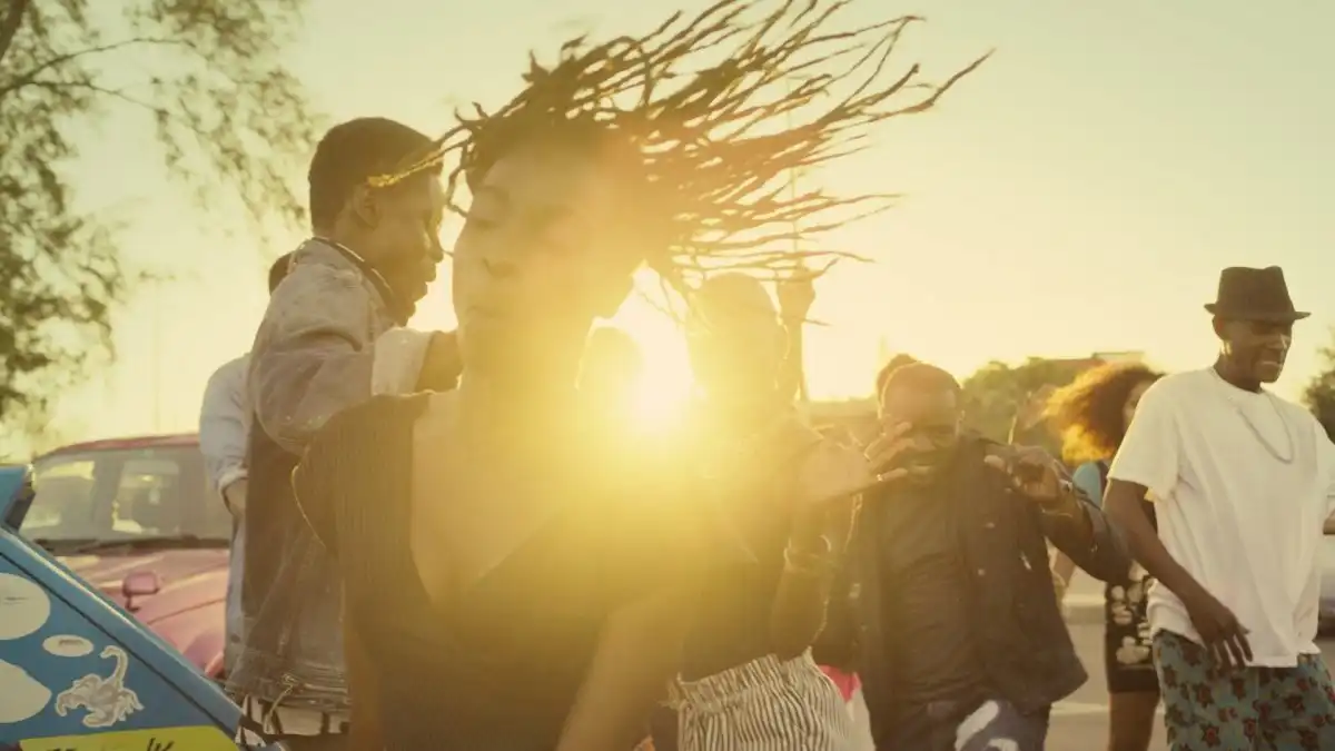 People dancing energetically outdoors at sunset, with sunlight streaming through.