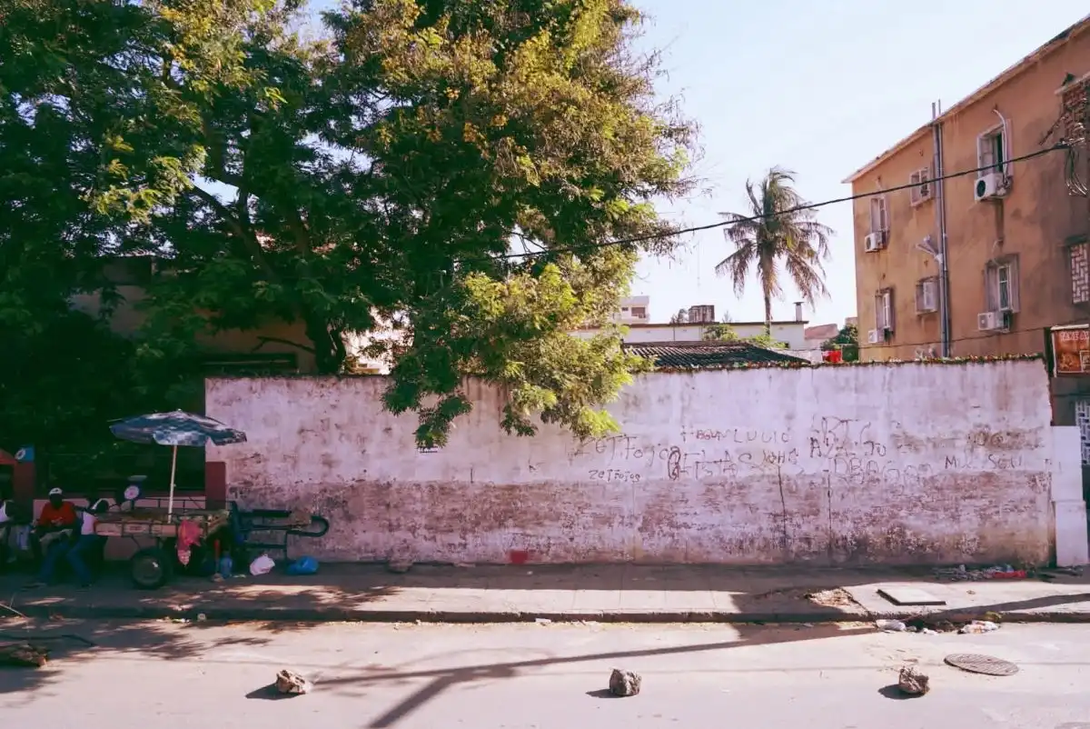 Street scene with a weathered wall, tree shade, and a small food cart on the left. Urban setting with a palm tree in the background.