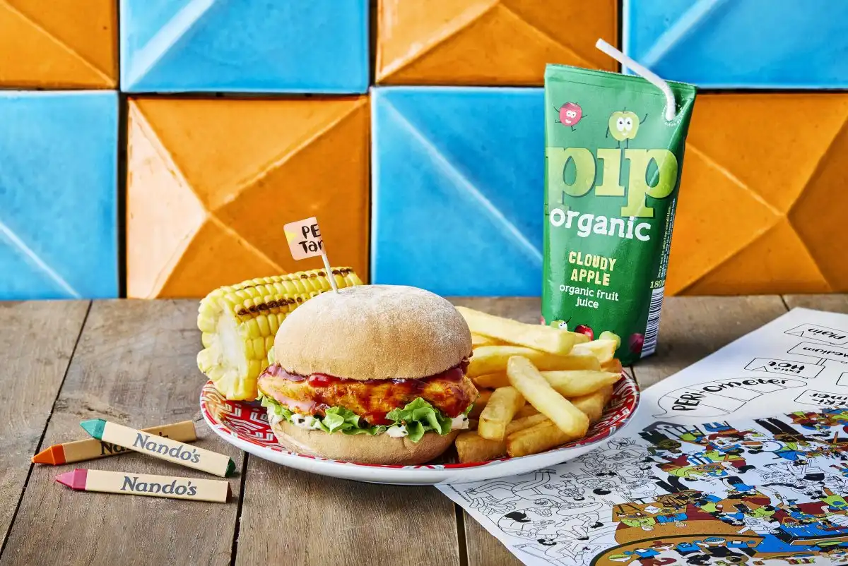 Nando's kid's meal with a chicken burger, corn, fries, Pip organic juice, and crayons on a colorful table.