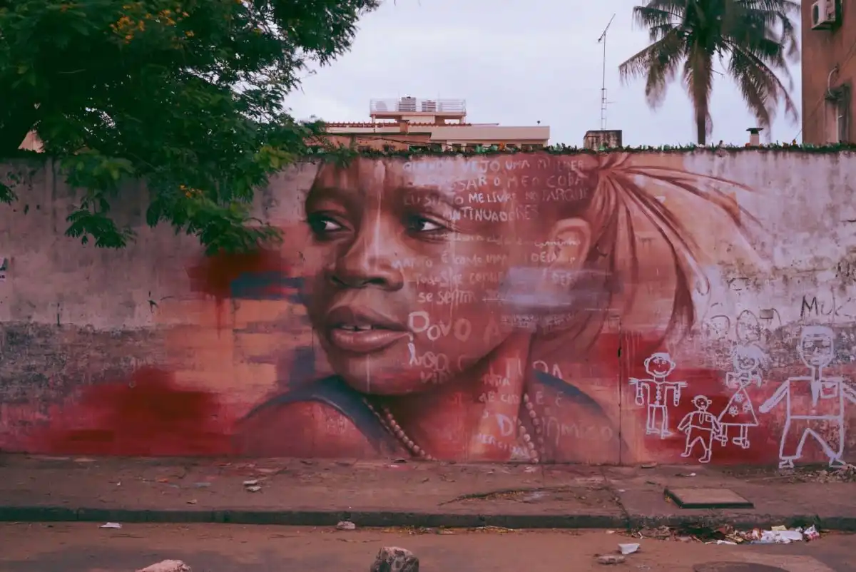 Street art of a woman's face on a wall, surrounded by graffiti and trees, with a palm tree in the background.