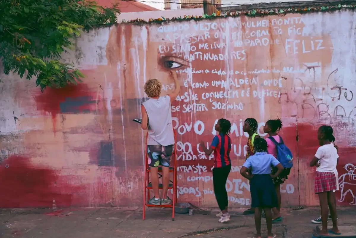 A person paints a mural on a wall while a group of children watch. The mural features large text and a detailed eye.