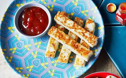 Grilled halloumi sticks on a patterned plate with a side of red dipping sauce.