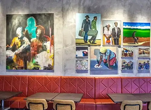 Restaurant wall adorned with vibrant, diverse artwork above red seating and wooden tables.