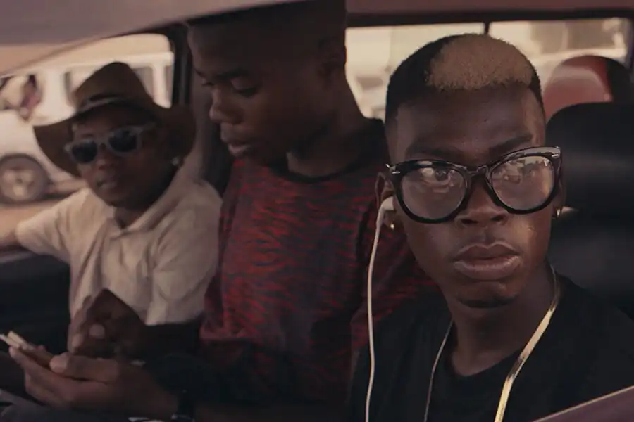 Three people sit in a car, one wearing large glasses and earphones, another in a hat, and the third in a patterned shirt.