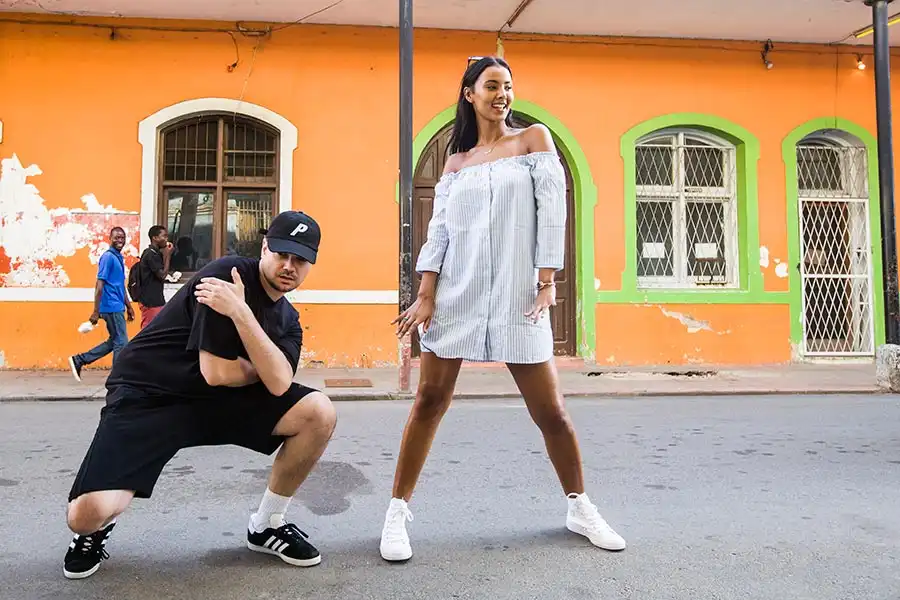 Two people pose confidently on a street with vibrant orange and green building in the background.
