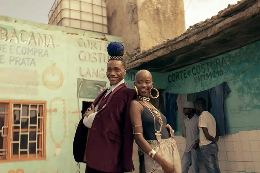 Two stylish individuals pose back-to-back, smiling in front of colorful, rustic buildings with vibrant murals and signage.