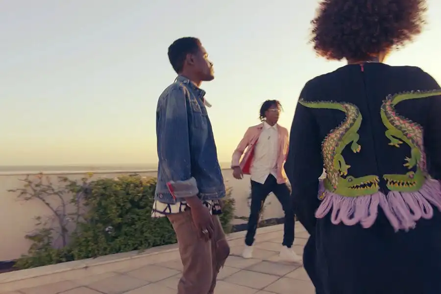 Three people walking and talking on a rooftop terrace at sunset, with one wearing a jacket featuring dragon designs.