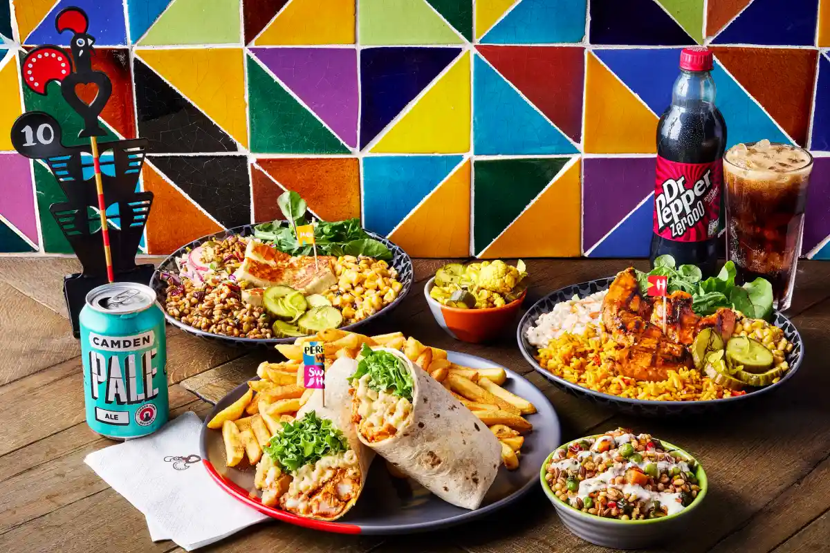 A colorful spread of various dishes including wraps, fries, salads, and drinks on a wooden table with a vibrant tiled background.