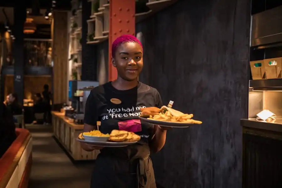 Server with pink hair holding plates of food, including fries and rice, in a dimly lit restaurant.