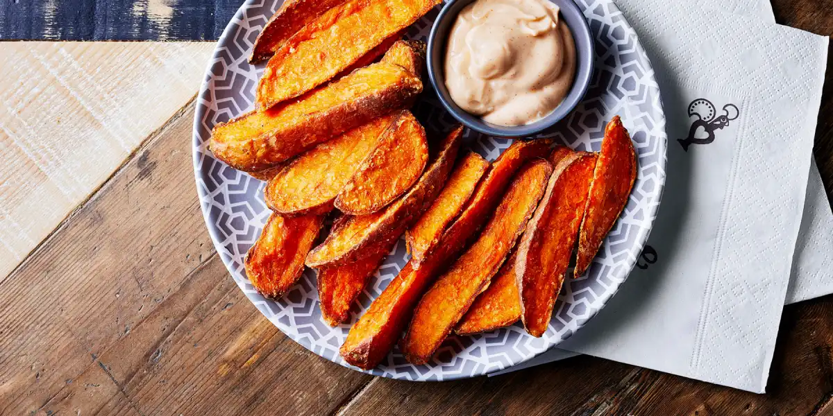 Plate of crispy sweet potato wedges with a creamy dip on a patterned plate, served on a wooden table.