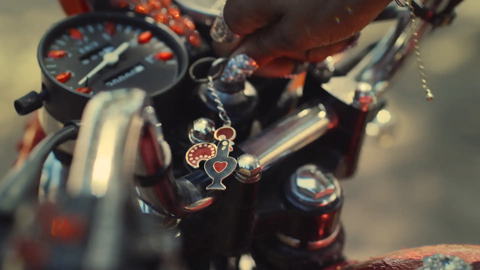 Hand holding a Nando's rooster keychain on a motorbike handlebar with a speedometer in the background.