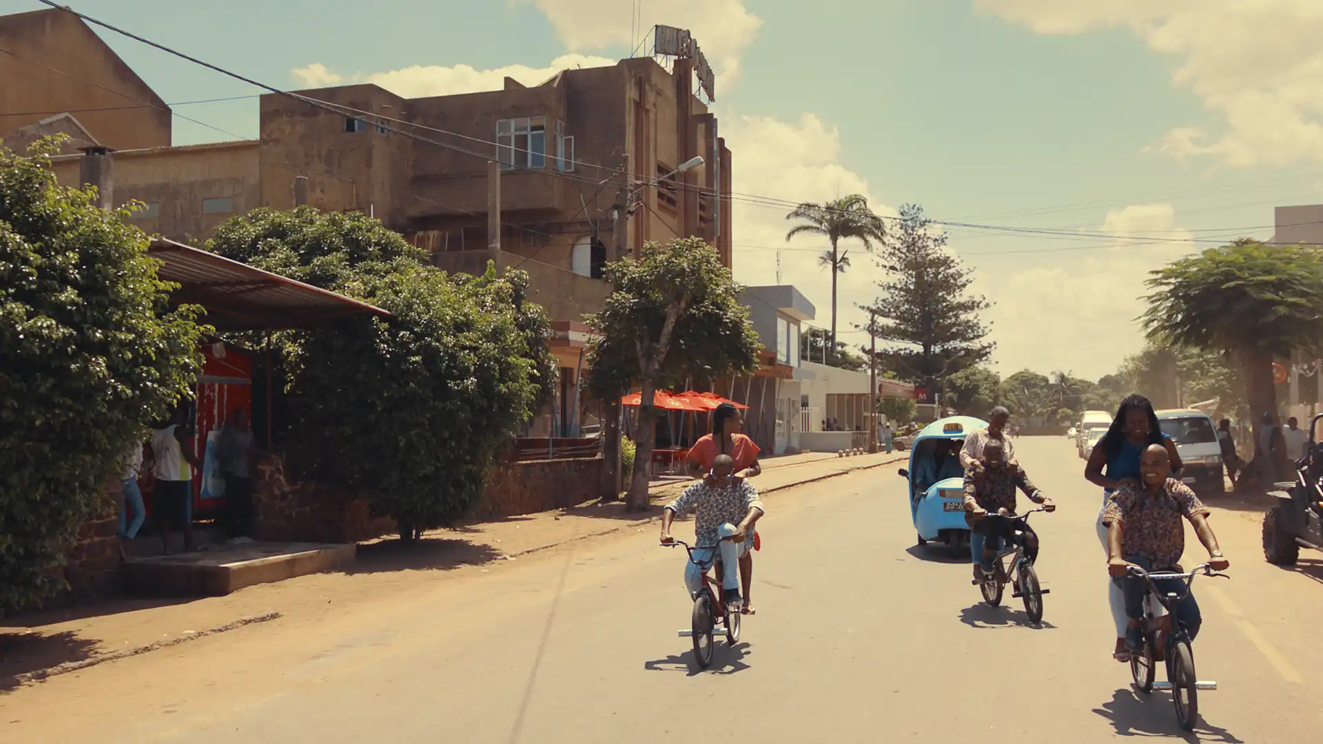 People riding bicycles and a tuk-tuk on a sunny street lined with trees and buildings.