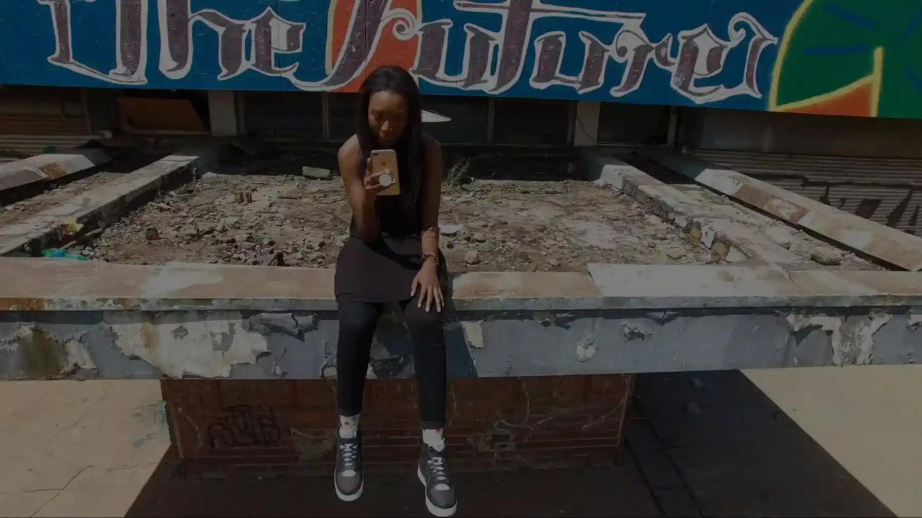 A person sits on a ledge, looking at their phone, with a colorful graffiti mural in the background.
